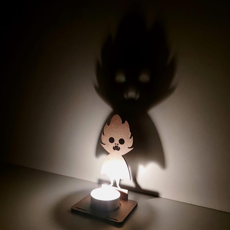 May include: A wooden candle holder with a cartoon ghost design. The candle holder casts a shadow of a larger ghost figure on the wall. The candle is lit, creating a warm glow and enhancing the shadow effect.