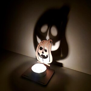 May include: A wooden pumpkin-shaped candle holder with a lit tea light. The candle's light casts a large pumpkin shadow on the wall. The pumpkin has cut-out eyes, nose, and a smiling mouth. A Halloween decoration.