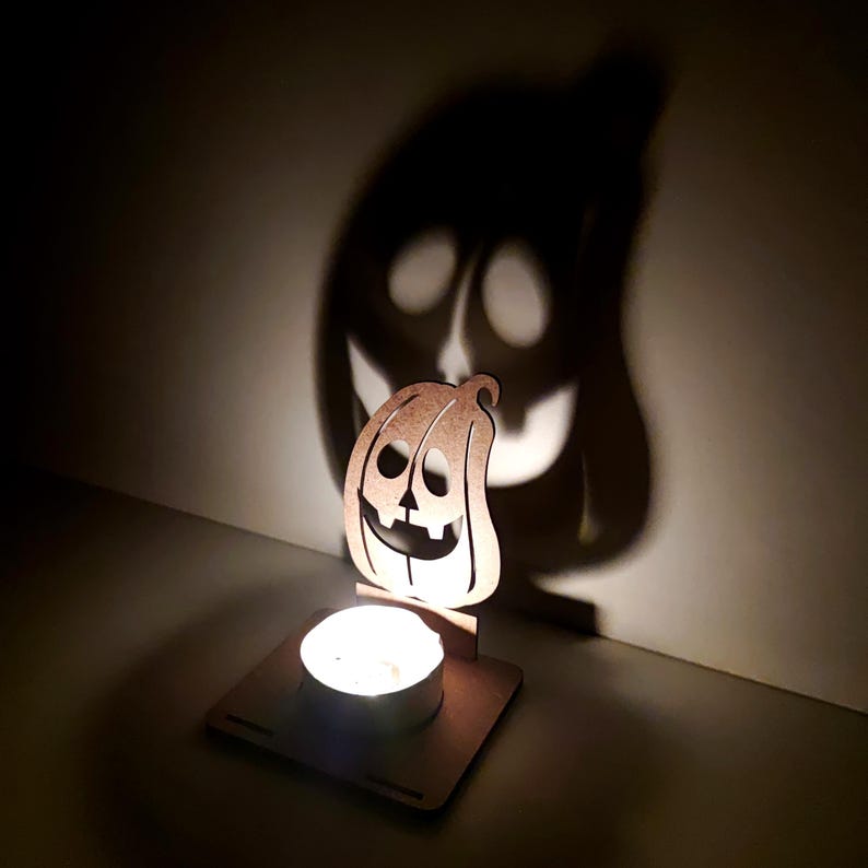 May include: A wooden pumpkin-shaped candle holder with a lit tea light. The candle's light casts a large pumpkin shadow on the wall. The pumpkin has cut-out eyes, nose, and a smiling mouth. A Halloween decoration.
