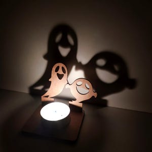 May include: A wooden Halloween-themed candle holder featuring two smiling ghost figures. The candle holder casts large ghost shadows on the wall behind it. The candle is lit, creating a warm glow.