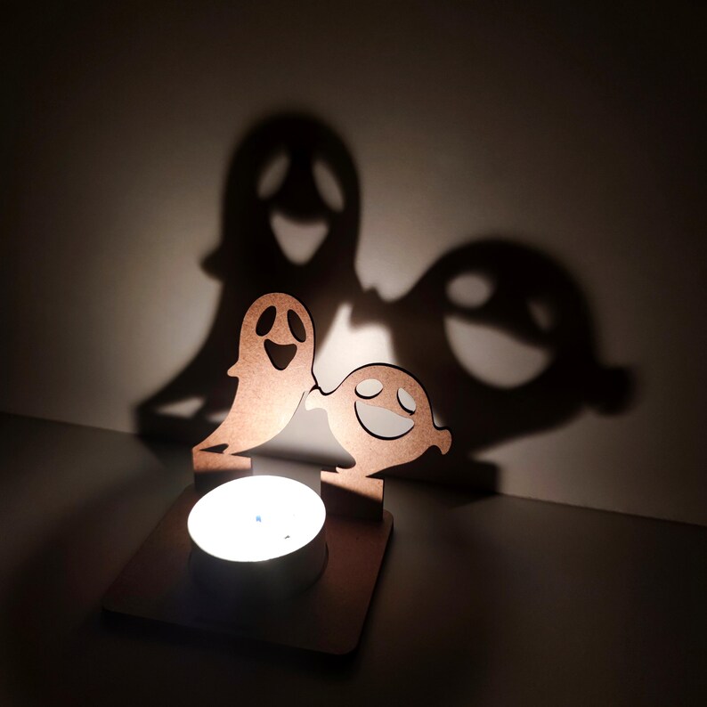 May include: A wooden Halloween-themed candle holder featuring two smiling ghost figures. The candle holder casts large ghost shadows on the wall behind it. The candle is lit, creating a warm glow.