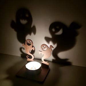 May include: A Halloween-themed candle holder with two wooden ghost cutouts casting large shadows on the wall. A lit tealight candle sits in the center, creating a spooky ambiance. The wooden cutouts have smiling faces.
