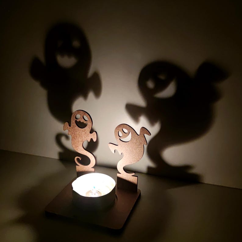 May include: A Halloween-themed candle holder with two wooden ghost cutouts casting large shadows on the wall. A lit tealight candle sits in the center, creating a spooky ambiance. The wooden cutouts have smiling faces.
