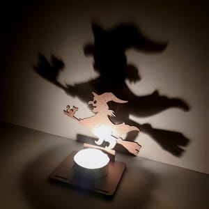 May include: A wooden witch silhouette on a broom, illuminated by a candle, casting a large shadow on the wall. The witch is wearing a pointed hat and holding a small object. The candle sits in a square wooden base, creating a Halloween-themed decoration.