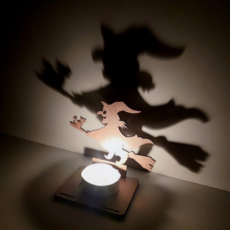 May include: A wooden witch silhouette on a broom, illuminated by a candle, casting a large shadow on the wall. The witch is wearing a pointed hat and holding a small object. The candle sits in a square wooden base, creating a Halloween-themed decoration.