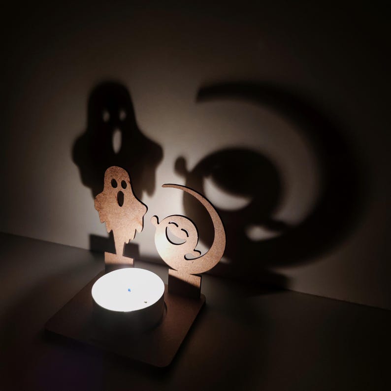 May include: A Halloween-themed candle holder made of wood. The holder features a ghost and a smiling moon design, casting shadows on the wall. A lit tealight candle sits in the center, creating a warm glow. The overall design is whimsical and festive.