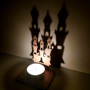 May include: A wooden castle-shaped candle holder with a lit tea light. The candle holder casts a shadow of a castle on the wall. The castle design has multiple towers and a square base. The color of the wood is brown.