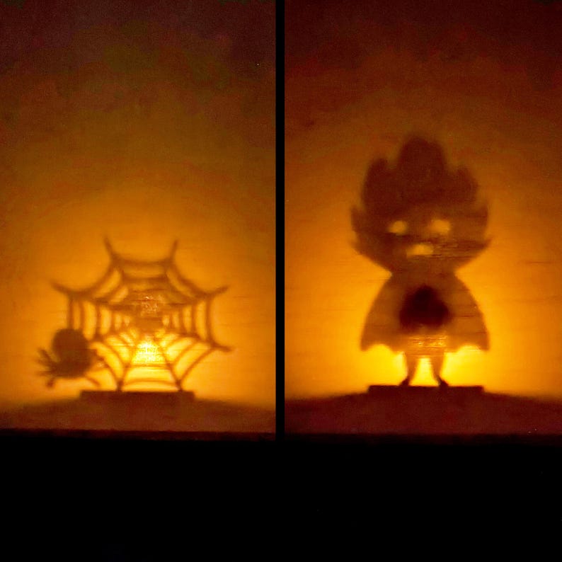 May include: Two Halloween-themed shadow light displays. One shows a spider and a web, the other a cartoon figure with a heart. The shadows are cast against a warm, orange background, creating a spooky ambiance.