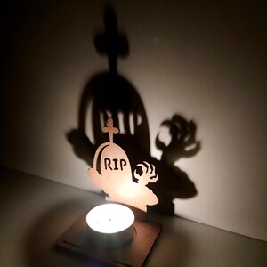 May include: A Halloween-themed candle holder featuring a wooden tombstone design with the letters "RIP" and a cross. The candle holder casts a shadow of a tombstone and a clawed hand on the wall. A lit tea light sits in front of the tombstone.