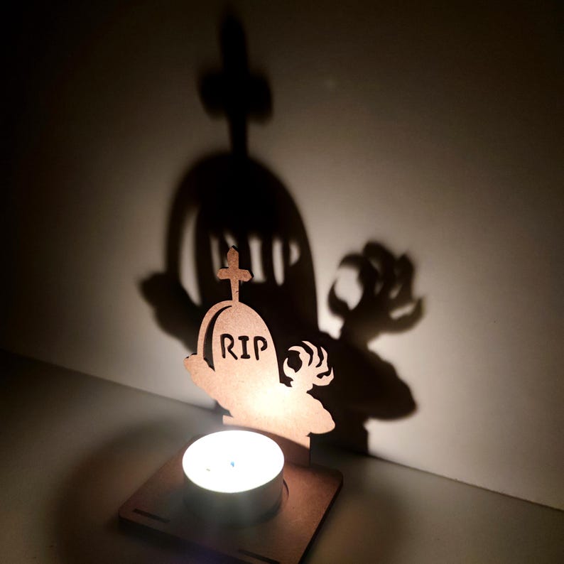 May include: A Halloween-themed candle holder featuring a wooden tombstone design with the letters "RIP" and a cross. The candle holder casts a shadow of a tombstone and a clawed hand on the wall. A lit tea light sits in front of the tombstone.