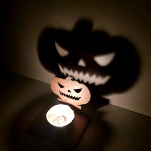 May include: A Halloween-themed candle holder with a carved pumpkin design. The candle holder casts a large shadow of a jack-o'-lantern face on the wall. The candle is lit, creating a warm glow.