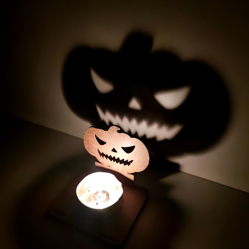 May include: A Halloween-themed candle holder with a carved pumpkin design. The candle holder casts a large shadow of a jack-o'-lantern face on the wall. The candle is lit, creating a warm glow.
