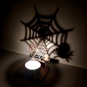 May include: A wooden Halloween-themed candle holder featuring a spider web and spider design. The candle holder casts a large shadow of a spider web and spider on the wall. The candle holder is made of light brown wood and holds a lit tea light candle.