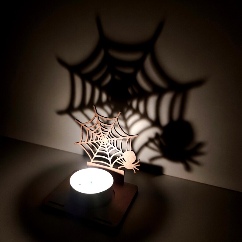 May include: A wooden Halloween-themed candle holder featuring a spider web and spider design. The candle holder casts a large shadow of a spider web and spider on the wall. The candle holder is made of light brown wood and holds a lit tea light candle.