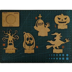 May include: A collection of unfinished wooden Halloween decorations, including a jack-o'-lantern, witch on a broom, spooky tree, and a tombstone with "RIP" carved into it. The pieces are displayed on a green cutting mat.
