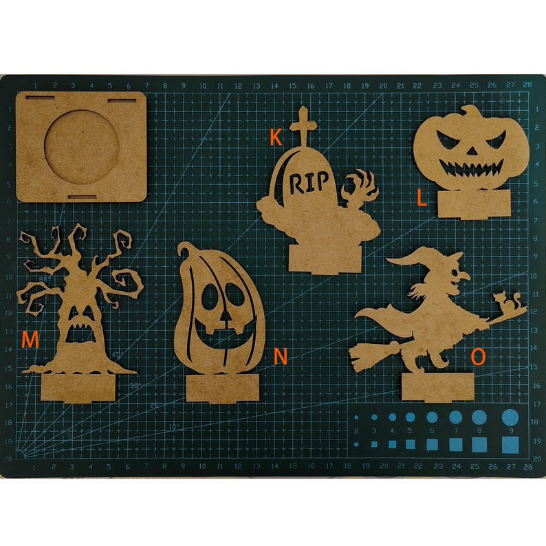 May include: A collection of unfinished wooden Halloween decorations, including a jack-o'-lantern, witch on a broom, spooky tree, and a tombstone with "RIP" carved into it. The pieces are displayed on a green cutting mat.