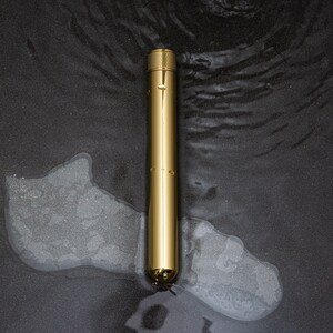 Puede incluir: Un bol&iacute;grafo de metal dorado con una parte superior texturizada y un cuerpo cil&iacute;ndrico liso. El bol&iacute;grafo est&aacute; sobre una superficie gris oscuro con gotas de agua.