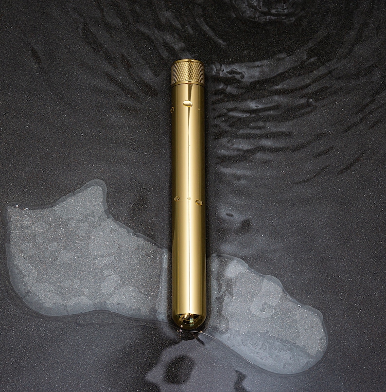 Puede incluir: Un bol&iacute;grafo de metal dorado con una parte superior texturizada y un cuerpo cil&iacute;ndrico liso. El bol&iacute;grafo est&aacute; sobre una superficie gris oscuro con gotas de agua.