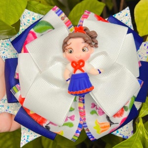May include: A white, blue, and multi-colored bow with a small doll figure in the center. The doll has brown hair, brown eyes, and is wearing a blue dress with a red bow.