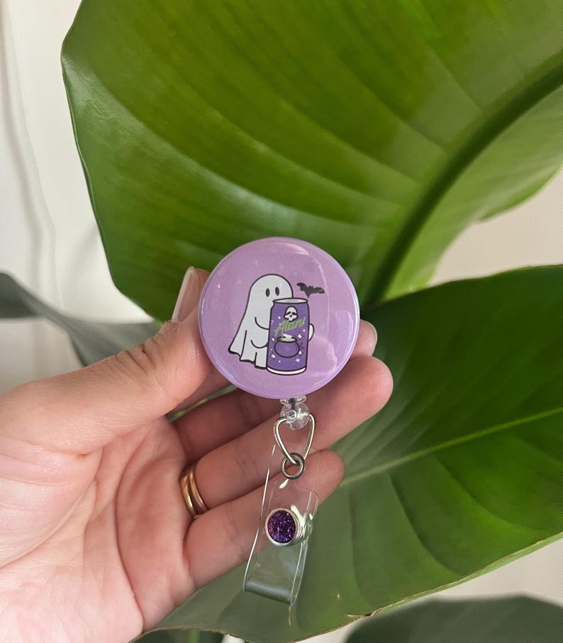 May include: A lavender badge reel featuring a cartoon ghost holding a purple can labelled "Mana". A black bat is above the can. The reel has a clear plastic strap and a purple glitter accent.