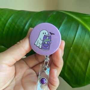 May include: A purple Halloween-themed badge reel featuring a ghost and a bat illustration. The badge reel has a clear plastic strap and a purple glitter accent. The ghost is holding a can that says "Alan!"