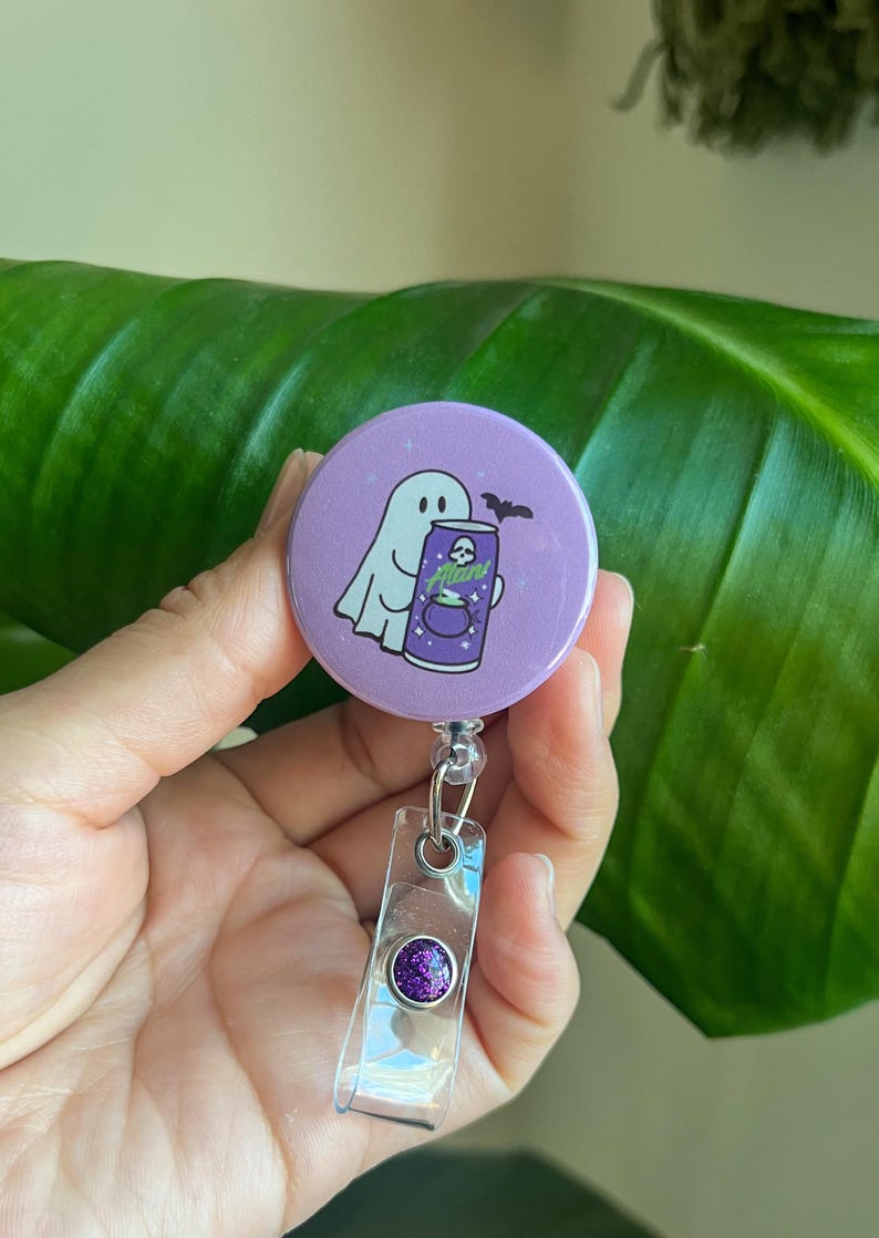 May include: A purple Halloween-themed badge reel featuring a ghost and a bat illustration. The badge reel has a clear plastic strap and a purple glitter accent. The ghost is holding a can that says "Alan!"