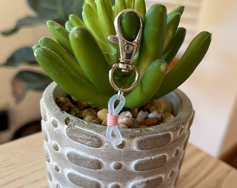 May include: A small succulent plant in a gray concrete pot with a silver keychain attached. The keychain has a pink and white rubber band in the shape of an infinity symbol.