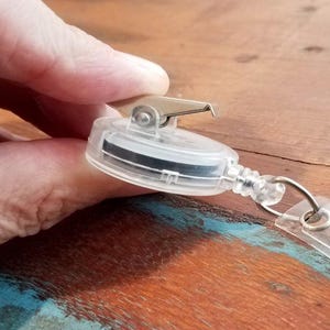 May include: Clear plastic retractable badge reel with a metal clip and a black band inside. The reel has a clear plastic cord and a metal ring for attaching a badge. The reel is held by a hand.