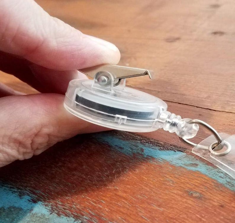 May include: Clear plastic retractable badge reel with a metal clip and a black band inside. The reel has a clear plastic cord and a metal ring for attaching a badge. The reel is held by a hand.