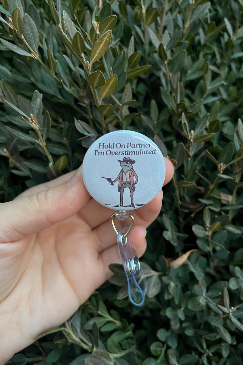 May include: A white badge reel with the text "Hold On Partner I'm Overstimulated" and a cartoon frog wearing a cowboy hat and holding a gun. The badge reel has a clear plastic strap and a metal clip.