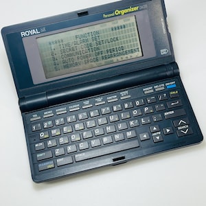 May include: A dark blue Royal 64K Personal Organizer DM200 with a small keyboard and a display screen. The screen shows a menu with options like Time/Alarm Set and Secret Code Set/Lock. The device has a contrast adjustment.