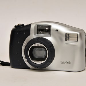 Op de afbeelding: Zilveren en zwarte Kodak digitale camera met een 28-50mm lens. De camera heeft een ingebouwde flitser en een polsband. Het Kodak-logo is zichtbaar aan de voorkant.