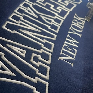 May include: A navy blue sweatshirt with white embroidered lettering that reads "YANKEES NEW YORK".