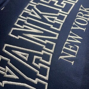 May include: A navy blue sweatshirt with white embroidered lettering that reads "YANKEES NEW YORK".