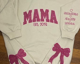 May include: A light beige sweatshirt with the word "MAMA" in pink glitter letters and the text "Est. 2019" below. The sleeves have pink glitter bows and the right sleeve has the names "Katherine", "Araceli", and "Sienna" written in pink glitter.