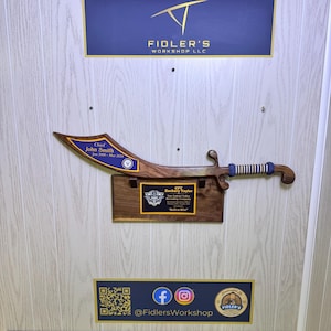May include: A wooden sword-shaped plaque with blue and gold accents, mounted on a light-colored wall. The plaque features engraved text, including names and dates, and is displayed above a smaller plaque. The business logo and social media handles are also visible.