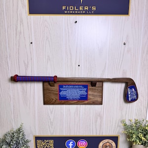 May include: A wooden golf club display featuring a golf club with a red and blue grip, mounted on a wooden plaque. The display includes a blue plaque with text and a logo, and is mounted on a white paneled wall.