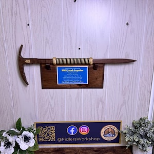 May include: A wooden ice axe with a dark brown handle and a metal pick. The handle is wrapped with a black and gold cord. A plaque with text is mounted below the axe. The text on the plaque reads "BM2 Jacob Logsdon".