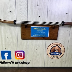 May include: A wooden walking stick with a black and white striped grip. The stick is mounted on a wooden plaque with a certificate for BM2 Jacob Logadon. The plaque is also decorated with a small wooden sign that says "Handcrafted By Fidler's Workshop, LLC Veteran Owned".