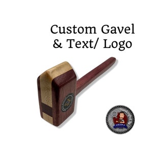 May include: A custom wooden gavel with a dark wood handle and a light wood head. The head has a black and gold emblem with a compass and square design. The gavel is handcrafted and has a silver tag with the text "Handcrafted by Fidler's Workshop".