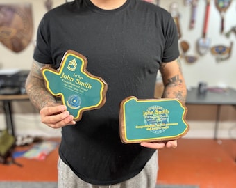 May include: Two wooden plaques with a green background and gold accents. The plaque on the left is shaped like the state of Texas and features the text "1st Sgt John Smith Texas National Guard Apr 2003 - June 2023". The plaque on the right is shaped like the state of Pennsylvania and features the text "John Smith PA National Guard Congratulations on your retirement and keep in touch".