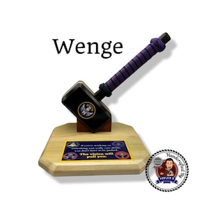 Puede incluir: Un martillo de madera hecho a mano con una cabeza negra y un mango envuelto en un cordón rojo, azul y negro. El martillo descansa sobre una base de madera con una cita y un logotipo. La palabra "Wenge" está encima del martillo.