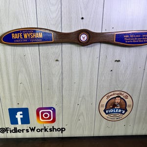 May include: A wooden airplane propeller with a blue and gold plaque that reads "CAPTAIN RAFE WYSHAM 22MAY1998 - 321JAN2025 NBK 19JUL2022 - 085EP2024 Thank you for your dedication to serve Naval Base Kitsap". The propeller is hanging on a wood panel wall. Below the propeller are social media icons for Facebook and Instagram with the text "@FidlersWorkshop". A round sticker with a cartoon man and the text "Handcrafted By FIDLER'S WORKSHOP LLC VETERAN OWNED" is on the wall.