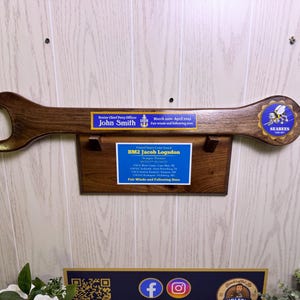 Puede incluir: Una placa de pared de madera oscura con forma de llave inglesa grande, que presenta una placa azul para John Smith, un emblema de los Seabees y una base rectangular con una placa azul para BM2 Jacob Logsdon, detallando su servicio y la frase Fair Winds and Following Seas.