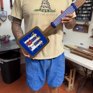 May include: A wooden plaque with a blue and white striped handle and a rectangular head. The head has a blue background with text that reads "MARSHALL K. BAILEY" and other details. The shirt in the background says "DON'T TREAD ON ME."