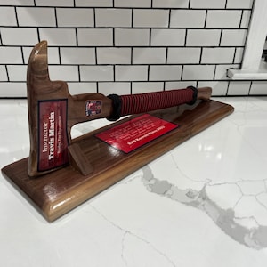 May include: A wooden firefighter's axe with a red and black striped handle is mounted on a wooden plaque. The plaque has a red label with the text "Instructor Travis Martin" and "RFD Recruit Class 2023".