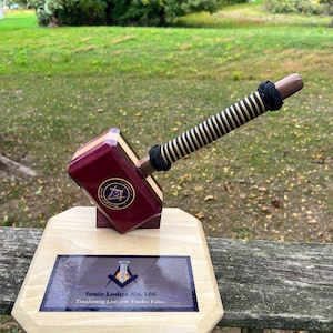 Puede incluir: Un mazo de madera con un mango de cuerda negro y dorado, descansando sobre una base de madera con una placa que dice "Ionic Lodge No. 185 Transforming Lives with Timeless Values".