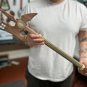 May include: A wooden halberd featuring a multi-pointed blade head with a circular emblem displaying the text United States of America. The handle is wrapped in a striped green and gold material, with light-colored cord bindings at both ends.