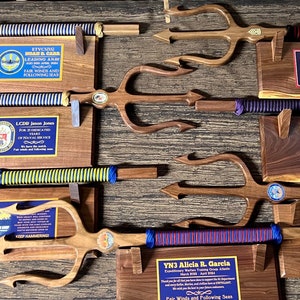 May include: Eight wooden tridents with blue, red, and yellow striped cords are mounted on wooden plaques. Each plaque has a name and a message. The plaques are on a wooden surface.