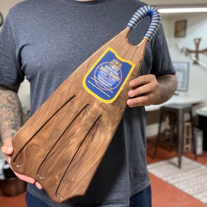 Può includere: Un vassoio da portata in legno a forma di pinna con un manico a nastro blu e oro. Il vassoio ha un'etichetta che dice "Sailor Jerry's John Smith's" e presenta un design nautico.